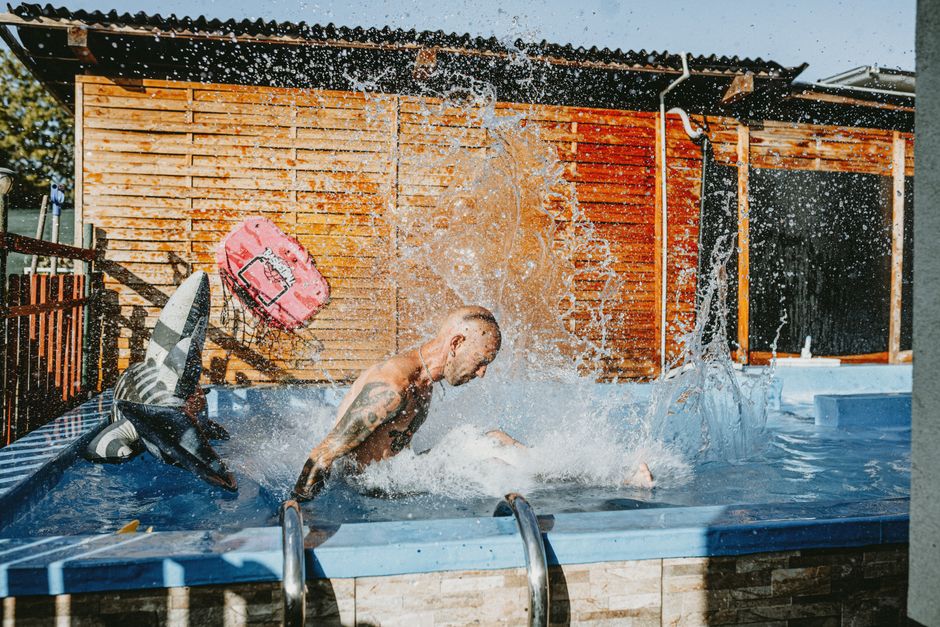 Un hombre tatuado chapoteando en la piscina de su jardín con un tiburón inflable. Diversión veraniega al aire libre.