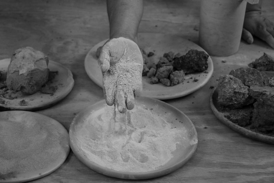 Imagen en blanco y negro de una mano tamizando polvo fino sobre platos con diversos materiales terrosos.
