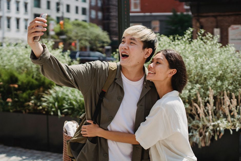 Una alegre pareja asiática se abraza y se hace un selfie en un entorno urbano al aire libre bajo la brillante luz del sol.