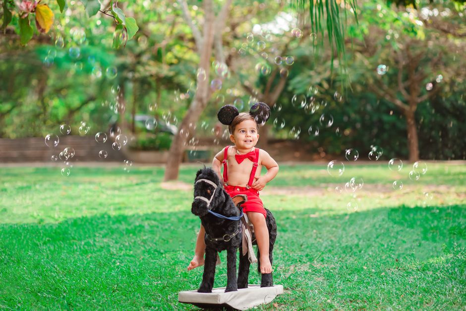 Un niño alegre con orejas de ratón se sienta en un caballo de juguete rodeado de burbujas en un parque soleado.