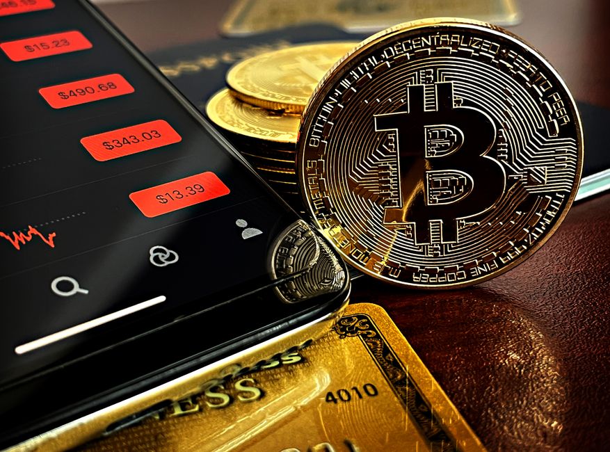 Una moneda de Bitcoin junto a la pantalla de un teléfono inteligente que muestra datos financieros, con tarjetas de oro al fondo.