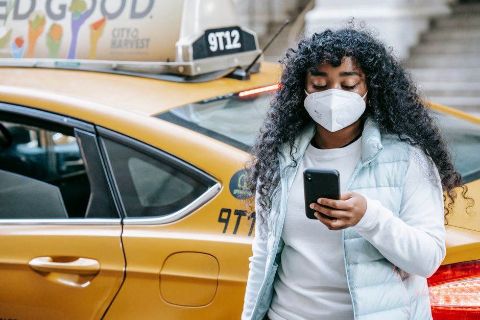 Joven con mascarilla usando un smartphone al lado de un taxi amarillo en un entorno urbano.