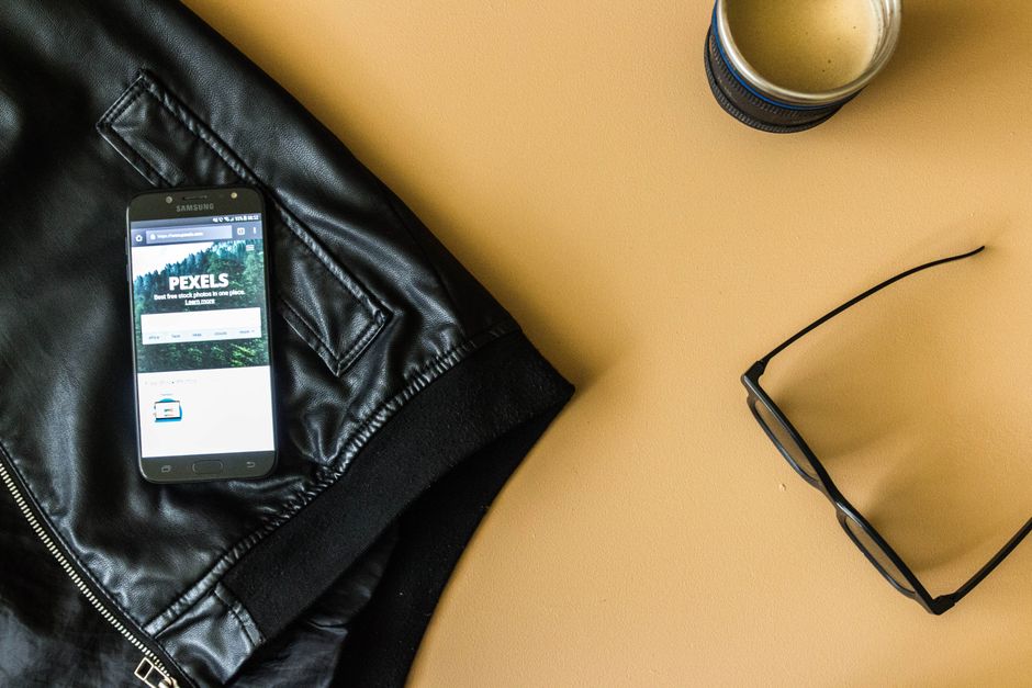 Disposición estilosa que presenta una chaqueta de cuero negro, un smartphone, café y gafas sobre una superficie beige.