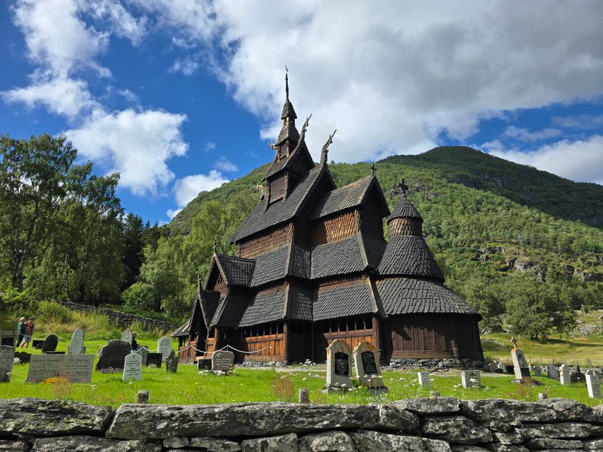 Histórica iglesia de madera noruega situada en un valle verde con lápidas en primer plano.