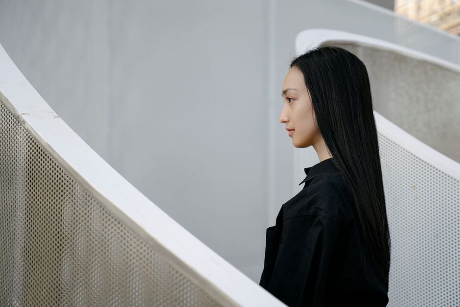 Una mujer con cabello largo en ropa negra posa elegantemente contra barandillas de escaleras modernas y curvas.