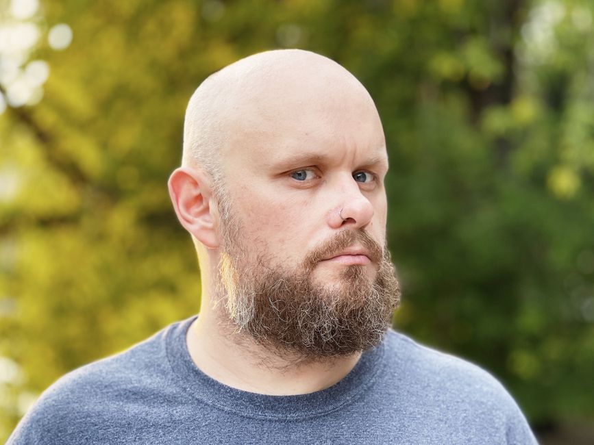 Retrato de un hombre calvo con barba, ojos azules y piercing en la nariz en un entorno natural al aire libre.