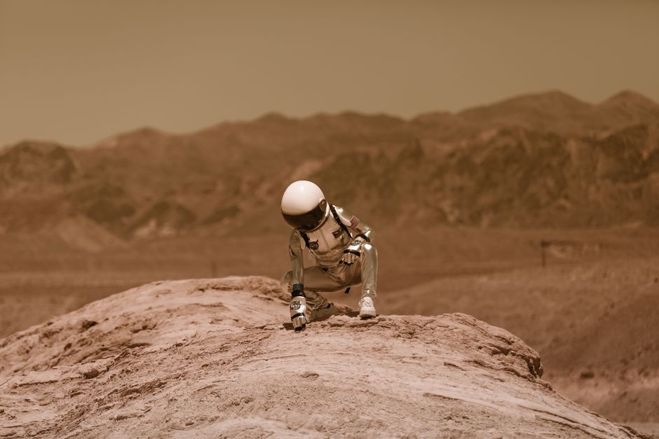 Un astronauta en un traje espacial explorando un entorno desértico similar a Marte, agachado sobre una superficie rocosa.