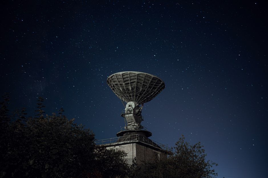 Una gran antena parabólica se erige frente a un fondo de un cielo nocturno lleno de estrellas, rodeada de vegetación.
