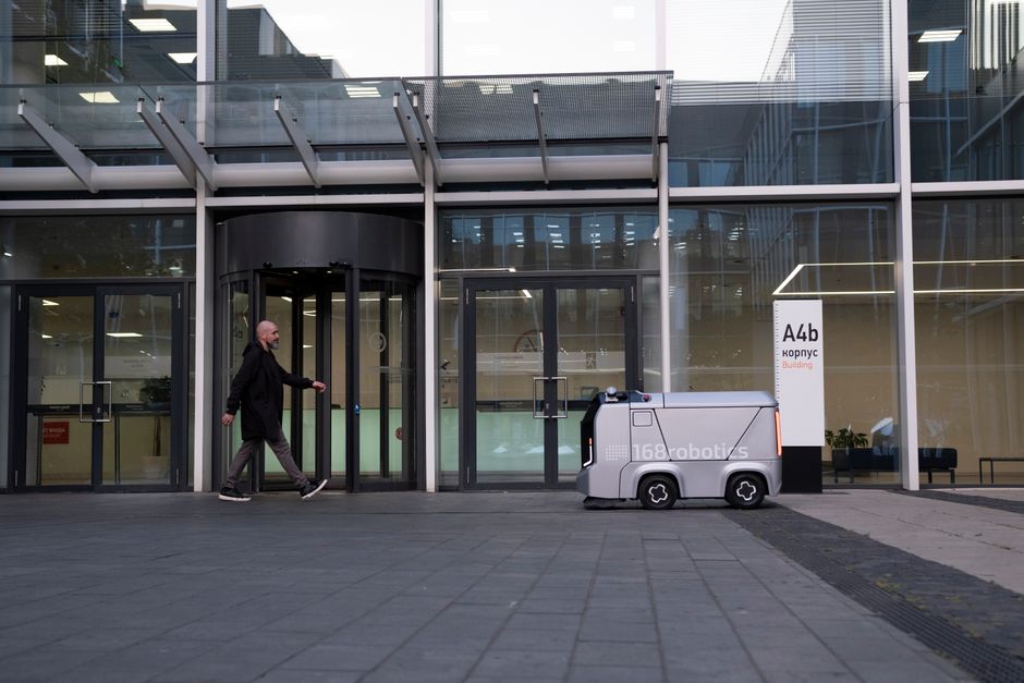 Un entorno de oficina moderno con un hombre y un robot de entrega autónomo fuera de un edificio cubierto de vidrio.