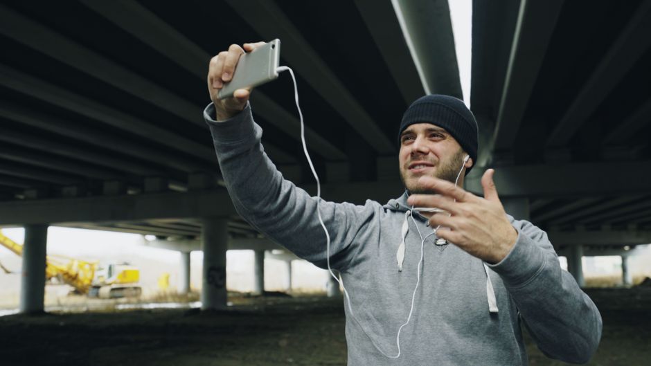 Hombre adulto toma un selfie bajo el puente, sonriendo con auriculares y vestido informal.