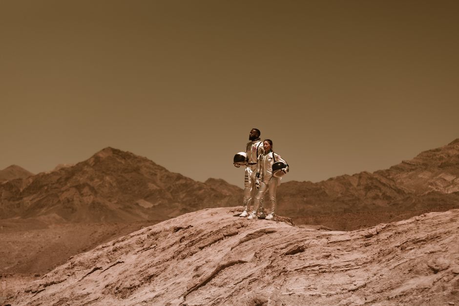 Dos personas en trajes espaciales están de pie en un paisaje árido, similar a Marte, bajo un cielo sepia.
