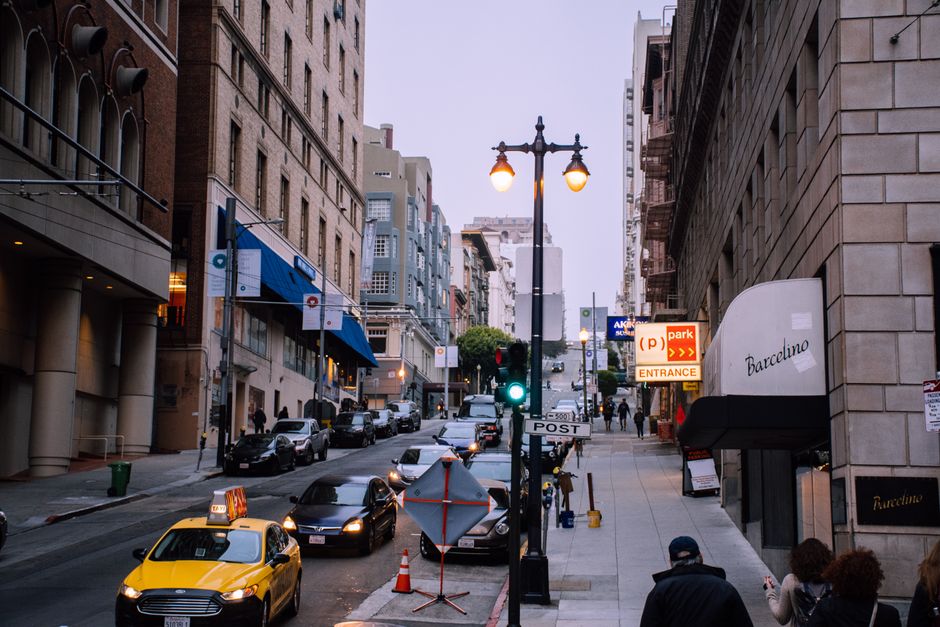 Una calle urbana concurrida con coches, peatones y tiendas al atardecer.